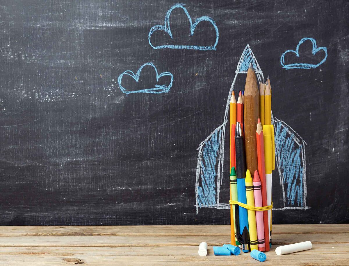 Eine schwarze Wandtafel, davor ein Holztisch mit einem Bündel Farbstifte, das vertikal aufgestellt wurde. Auf der Tafel sind dann mit Kreide die Umrisse einer Rakete gemalt, blaue Wolken drumherum.