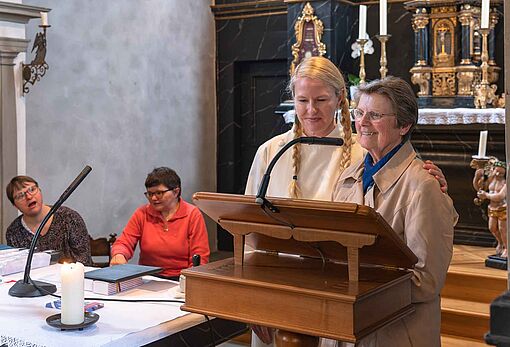 Rechts vorne im Bild zwei Frauen am Rednerinnenpult in der Kirche. Die Frau im weissen Talar hält ihren Arm um die Schulter der älteren Frau im hellbraunen Mantel. Fröhliche Gesichter. Links im Hintergrund sitzen noch zwei Menschen und hören bzw. sehen zu.