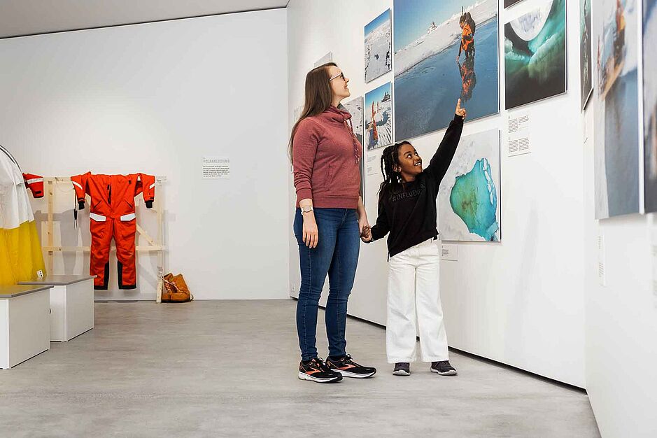 Eine junge Weisse Frau mit einem Schwarzen Mädchen in einem Ausstellungsraum. Sie stehen Hand in Hand, das Mädchen zeigt mit strahlendem Gesicht auf eine der Fotografien an der Wand.