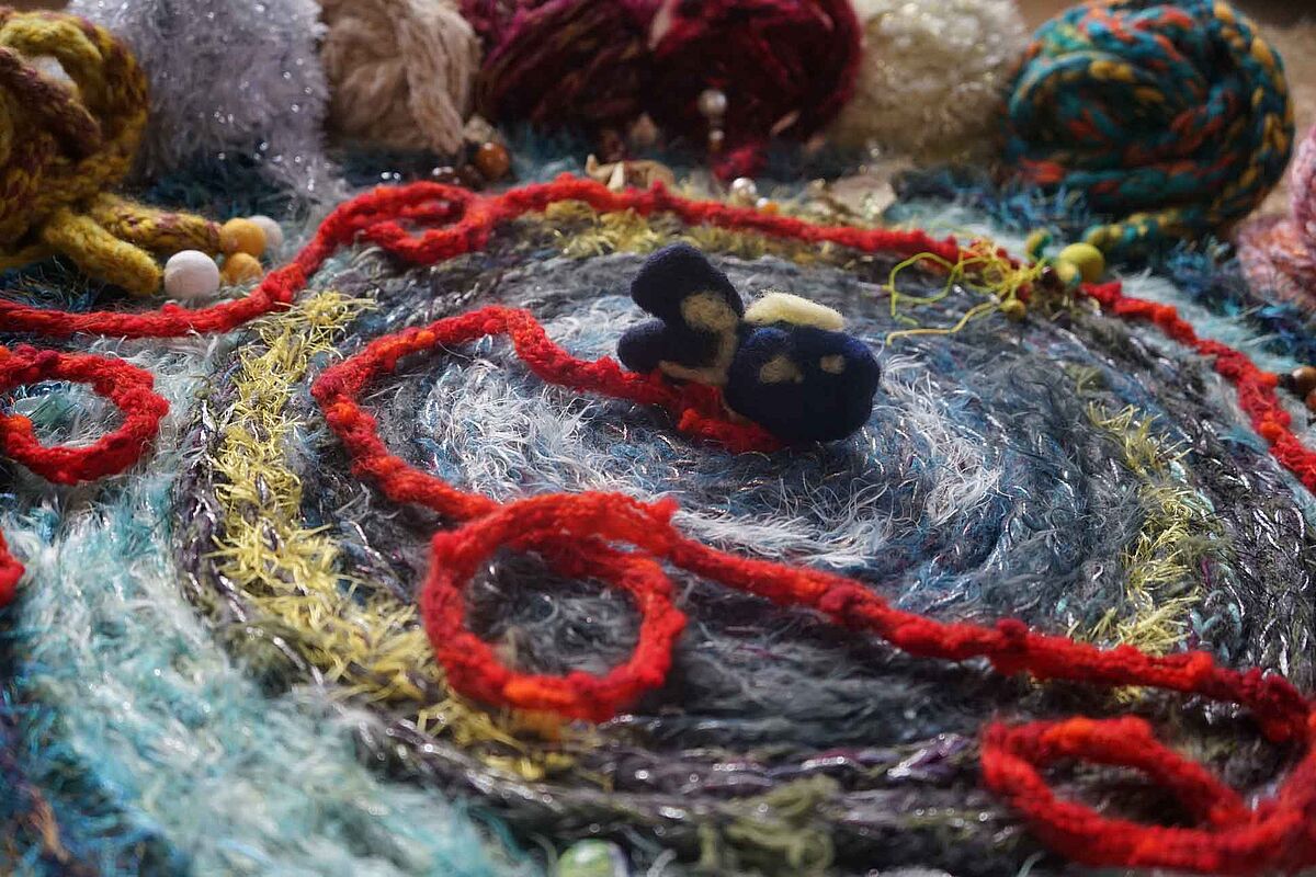 Eine rote Legeschnur aus Wolle liegt ausgebreitet auf einem Teppich, darum herum aufgerollte Legeschnüre als Knäuel.