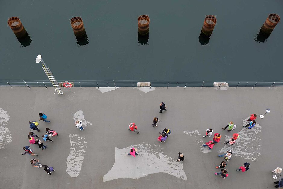 Das Bild ist aus der Vogelperspektive aufgenommen. Der untere Teil des Bildes ist ein Quai, auf dem viele Menschen laufen, der obere Teil des Bildes ist Wasser mit Pfeilern drin. Die Symmetrie des Fotos strahlt Ruhe aus.