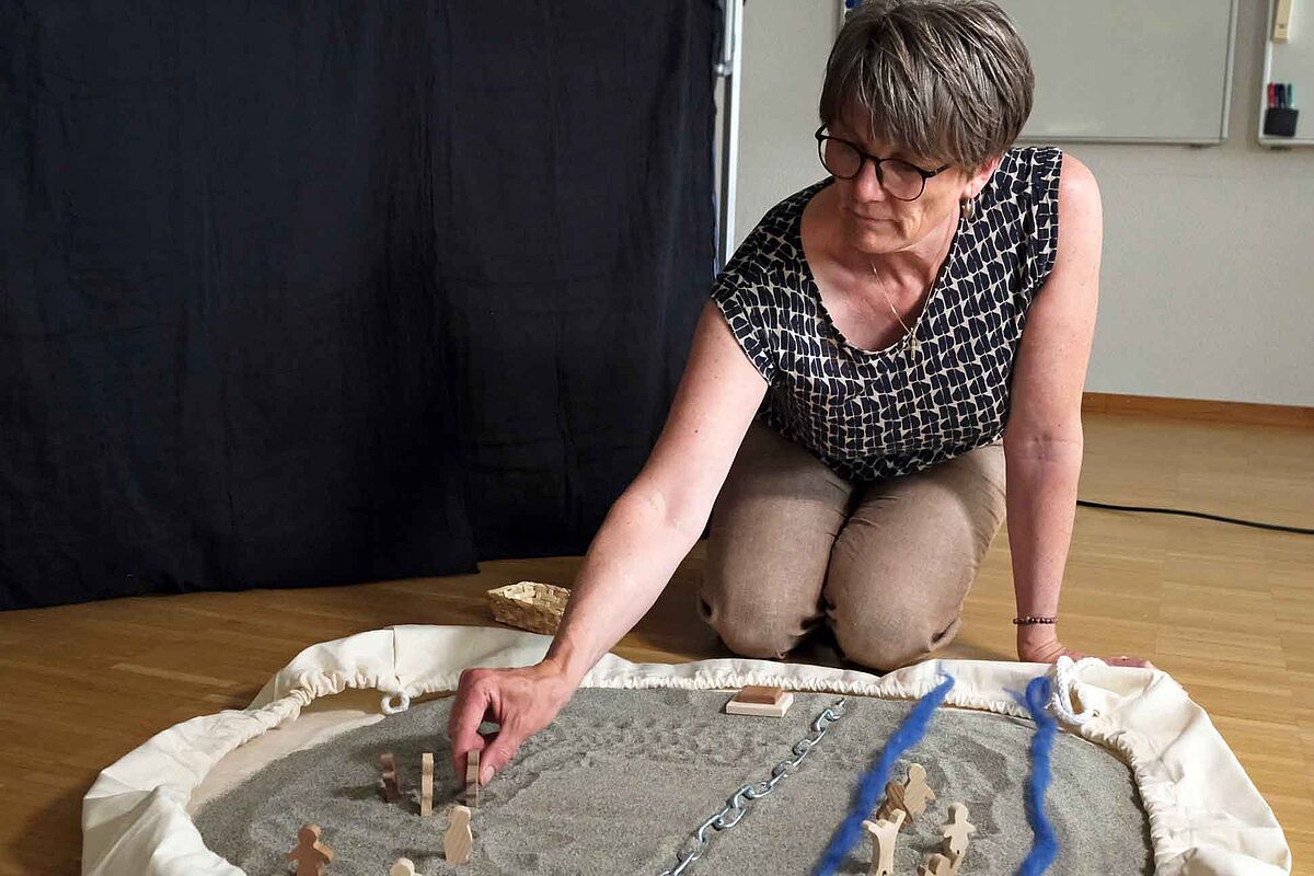 Eine Frau kniet hinter einem ausgebreiteten Sack, darauf ist Sand verteilt. Im Sand sind verschiedene Holzfiguren, eine Metallkette und zwei blaue Schnüre bewusst hingelegt, sodass eine Landschaft entstanden ist. Die Frau bewegt eine der Figuren im Sand. Sie ist sehr konzentriert.