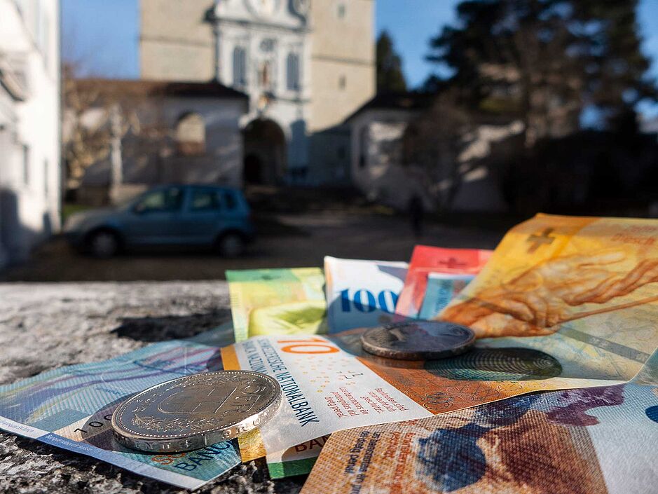 Im Vordergrund Geldnoten, die auf einer Mauer liegen, im Hintergrund die Hofkirche Luzern