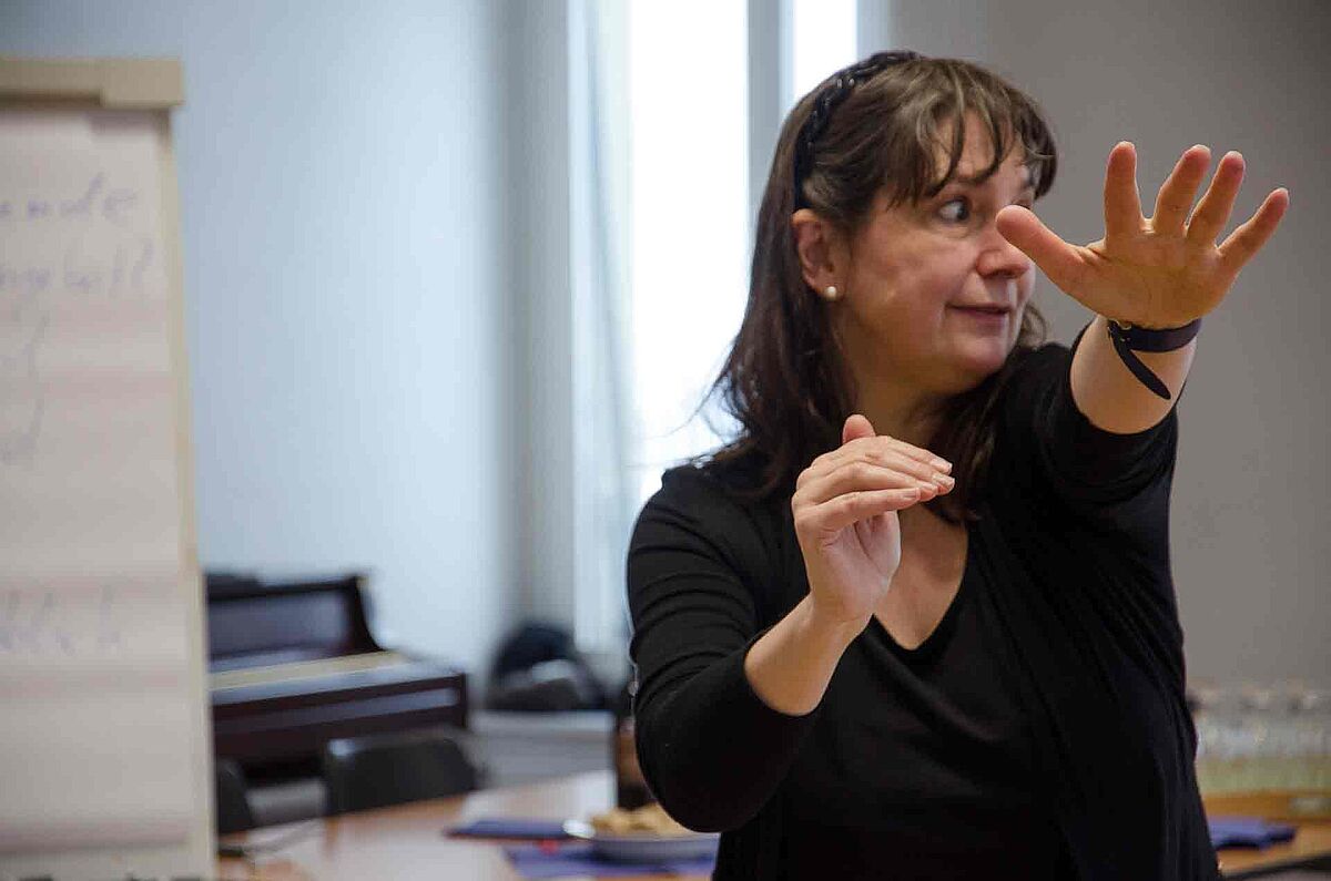 Eine Frau macht eine grosse Gebärde, ihre linke Hand ist gerade nach vorne ausgestreckt