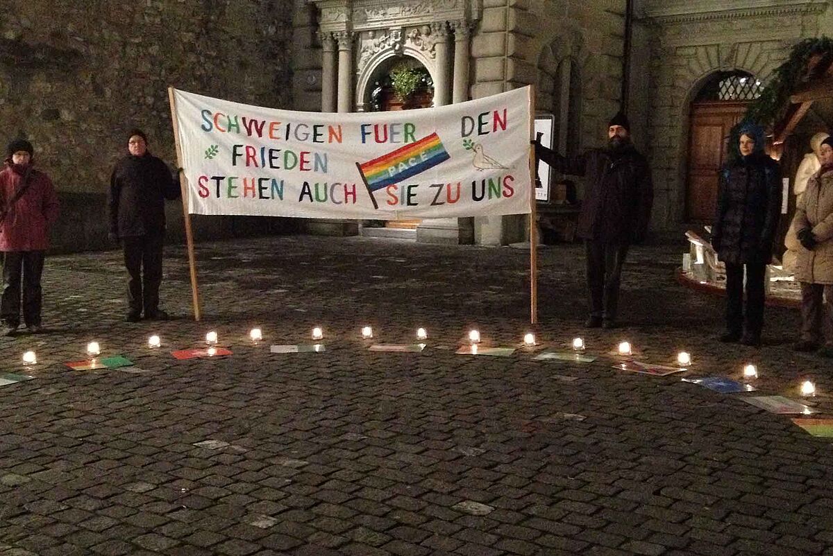 Es ist dunkel. Auf dem Bild ist ein angeschnittener Kreis aus Kerzen am Boden zu sehen, dahinter stehen Menschen, zwei halten ein grosses Transparent, auf dem in bunten Buchstaben steht: Schweigen für den Frieden. Stehen auch Sie zu uns.