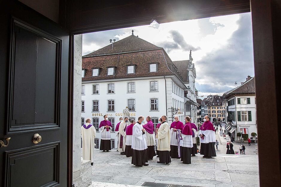 Blick durch eine Kirchentür nach draussen; auf dem Vorplatz stehen Priester